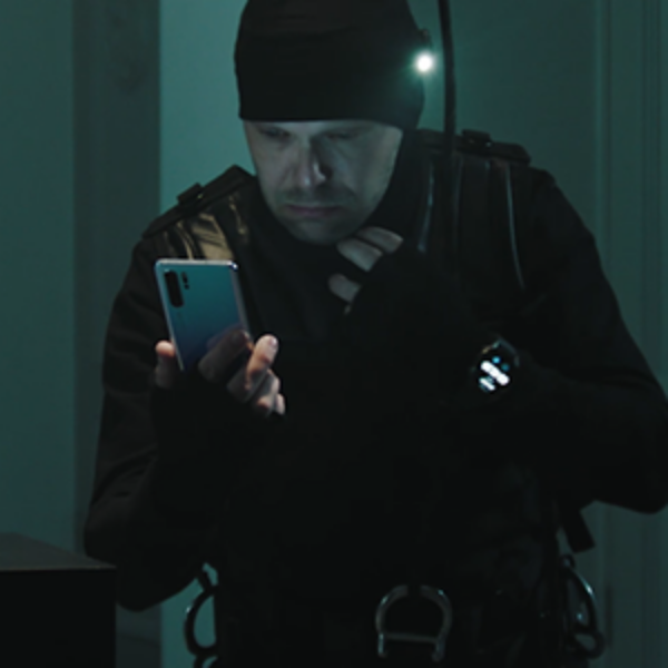 Ninja-asuun pukeutunut mies vetää maskin alaosan pois kasvoilta ja katsoo Huawei-puhelinta.