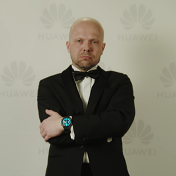 Vakavailmeinen mies puvussa seisoo kädet ristissä seinän edessä, jossa Huawein logoja.
