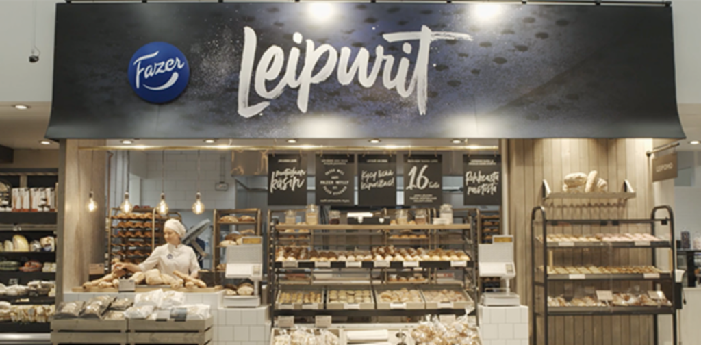 Fazerin myyntipiste, jonka hyllyt ovat täynnä leivontatuotteita ja tiskin takana valkopukuinen leipuri.