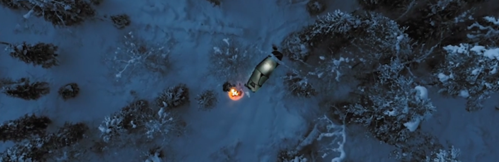 Lintuperspektiivi teltasta ja nuotiosta lumisen metsän keskellä. Nuotion ääressä istuu henkilö.