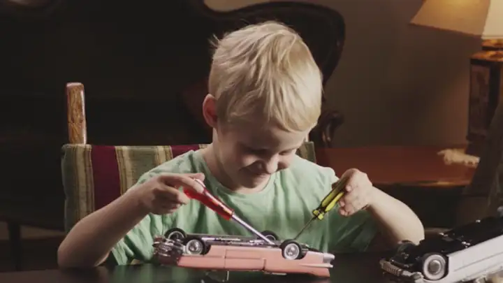 Hymyilevä poika korjailee leikkiautoa kahdella ruuvimeisselillä. Videolooppi.
