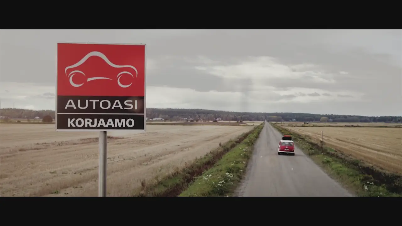 Laajakuva maisemasta, jossa punainen auto ajaa kohti horisonttia. Etualalla Autoasi korjaamo kyltti. Videolooppi.
