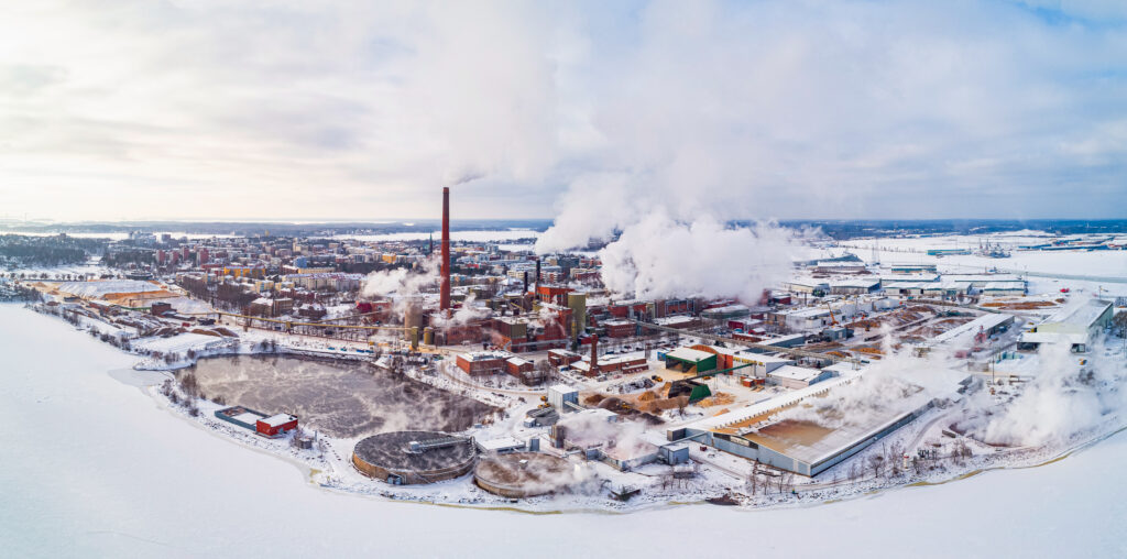 Laajakuva talvisesta maisemasta, josa suuri punainen tehdasalue, rantaa sekä kaupunkia. Tehtaan piipusita tulee savua.