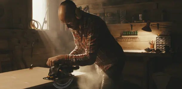 Mies sahaa puulevyä sähköisellä FXA-työkalulla. Pölyä lentää ilmaan. Videolooppi.