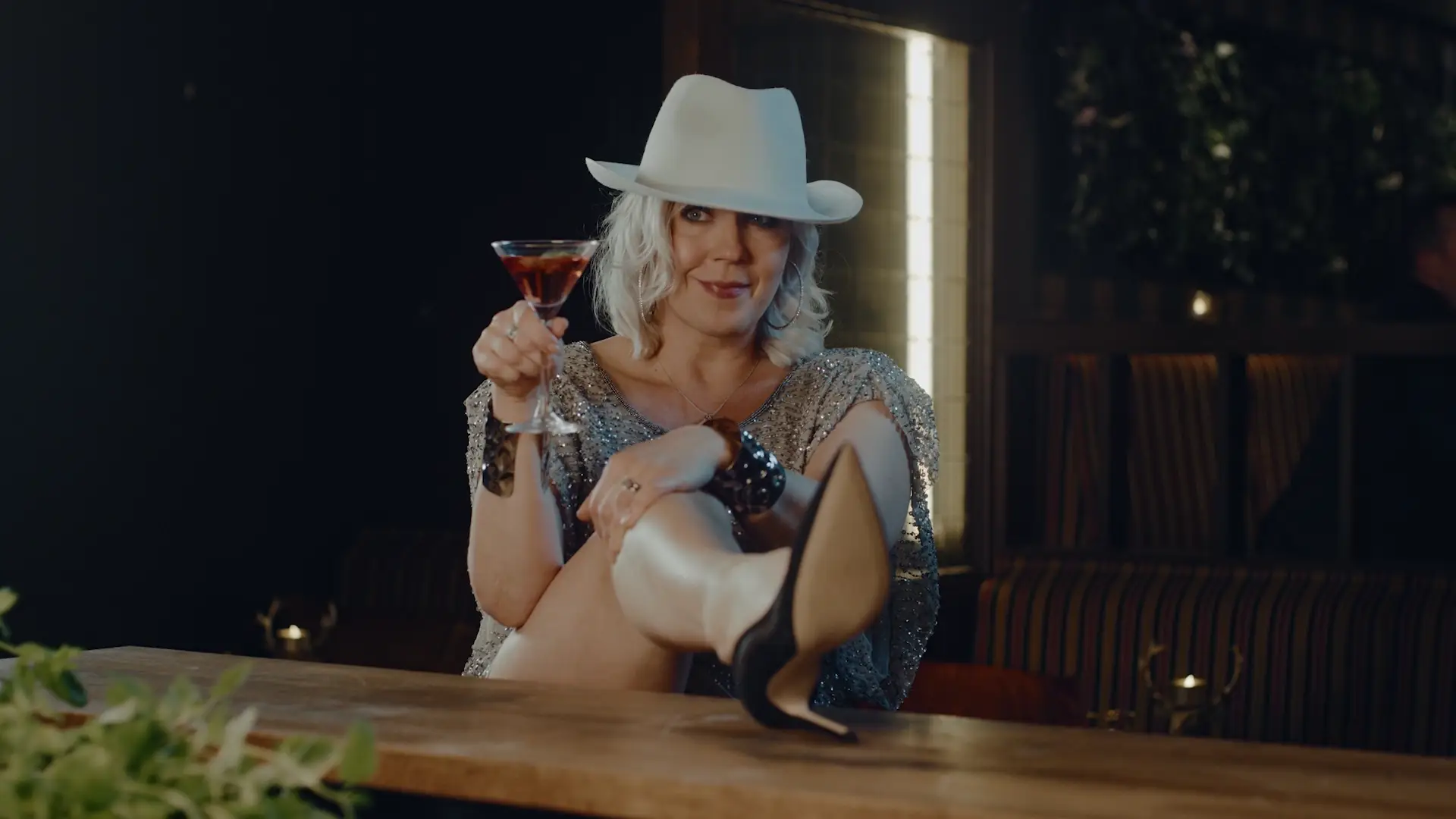 Vappu Pimiä valkoisessa cowboyhatussa istuu tuolilla jalat pöydällä. Pitelee drinkkilasia kädessä ja naurahtaa. Videolooppi.