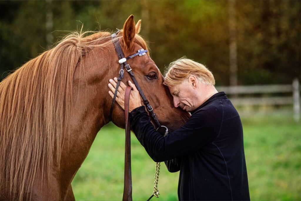 Sivuprofiili miehestä, joka nojaa kasvojaan ruskean hevosen turpaa vasten. Taustalla nurmikkoa ja metsää.