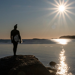 Henkilö märkäpuvussa seisoo kalliolla, meren äärellä surffilauta kainalossa. Taivaalla kirkas ilta-aurinko.