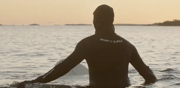 Mies märkäpuvussa, selkä katsojaan päin. Mies vedessä surffilaudan päällä. Videolooppi.
