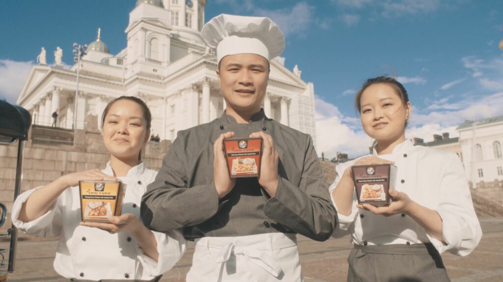 Mies ja kaksi naista seisoo Helsingin Tuomiokirkon edessä pidellen Kitchen Joy -ruokapakkauksia.