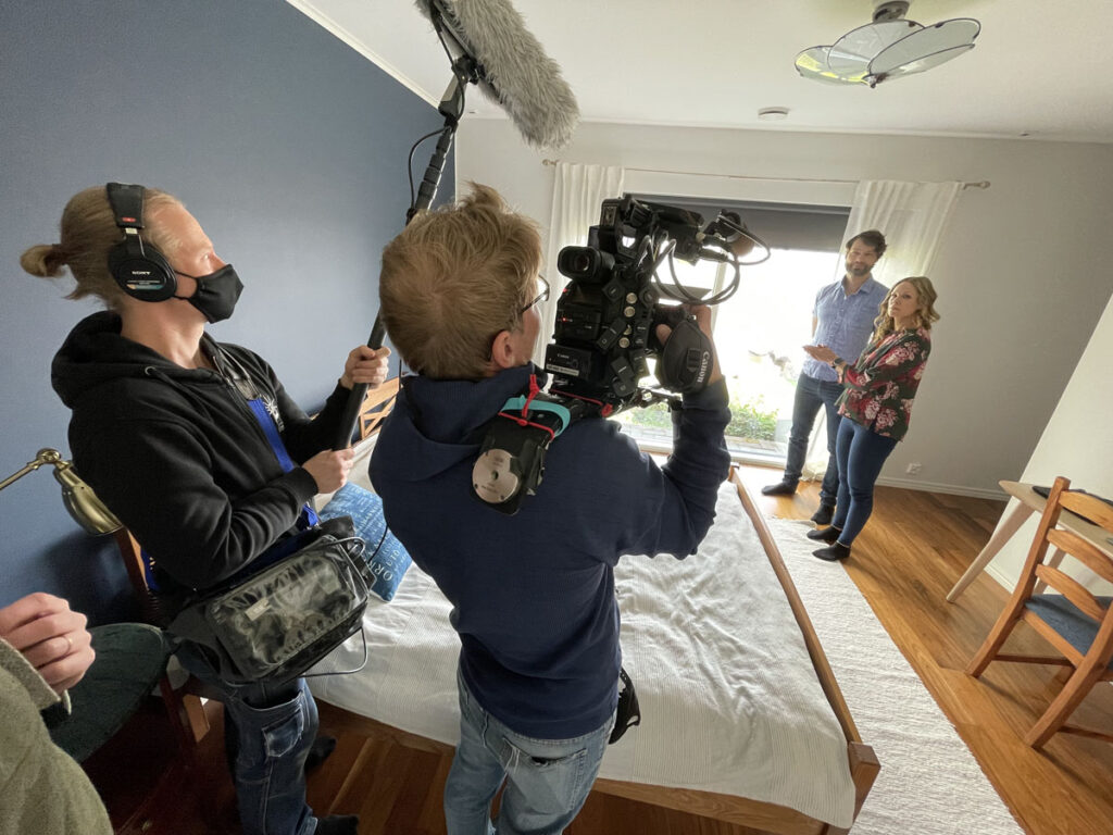 Behind the Scenes. Videokuvaaja ja äänittäjä työssään, kauempana mies ja nainen joita kuvataan.