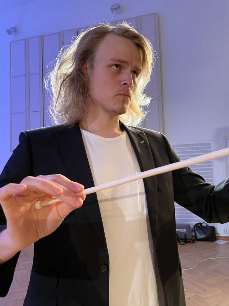 Lähikuva pitkähiuksisesta kapellimestarista, jolla on tahtipuikko kädessä.