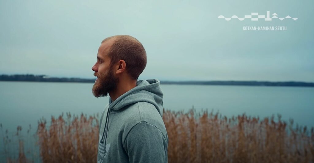 Teemu Pukki seisoo hupparissa meren äärellä, taustalla näkyy kaisloja, meri ja vastarannalla metsää.
