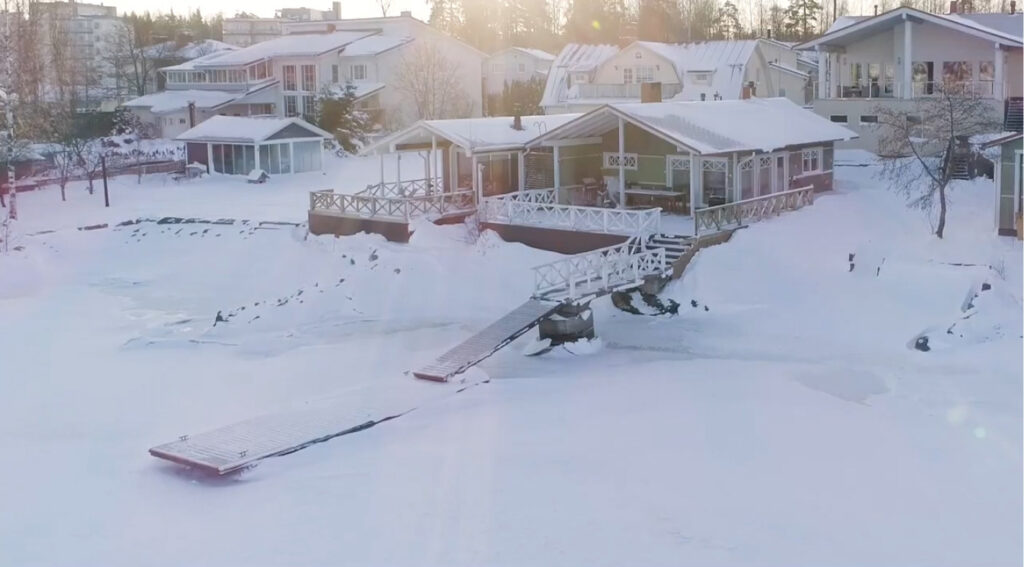 Talvinen rantamaisema, jossa lumen peittämä jää, laituri sekä rakennuksia aivan rannassa.