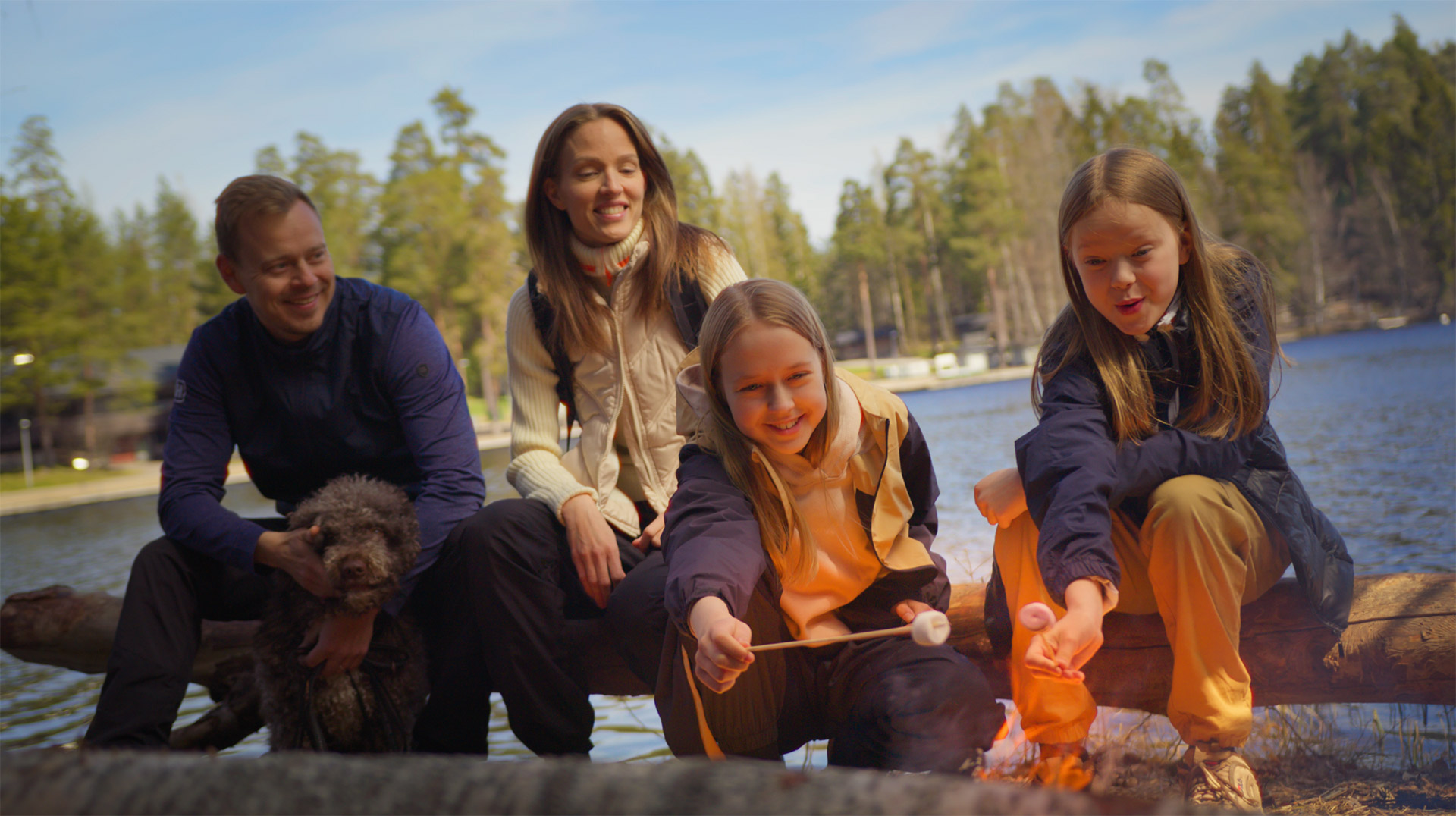 Mies, nainen ja kaksi tyttöä istuu veden äärellä ja grillaavat nuotiossa vaahtokarkkeja.